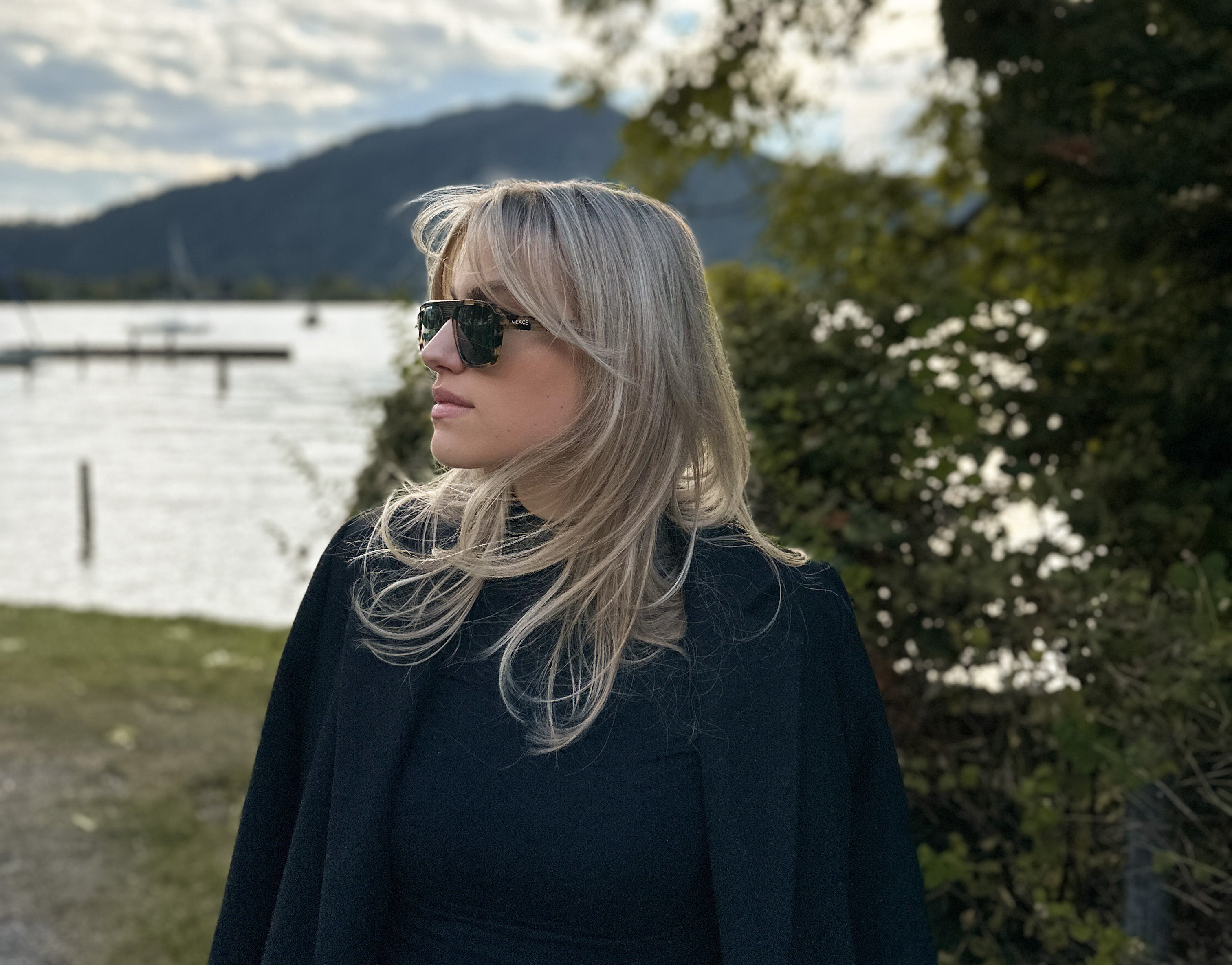 Eine elegante Frau spaziert am herbstlichen Seeufer entlang und trägt eine schicke CEACE Sonnenbrille. Die Sonnenbrille schützt vor blendendem Sonnenlicht und verleiht ihrem Look Eleganz.